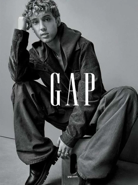 Monochrome portrait of a young adult male model in casual apparel sitting on a box with the logo 'GAP' dominating the foreground.
