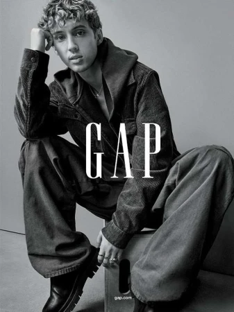 Monochrome portrait of a young adult male model in casual apparel sitting on a box with the logo 'GAP' dominating the foreground.