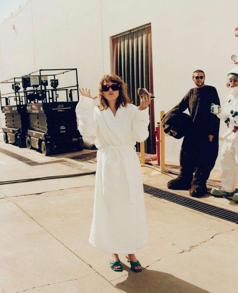 Woman in white robe poses with a bird, two people behind her in an industrial setting with aerial lifts and a closed door.