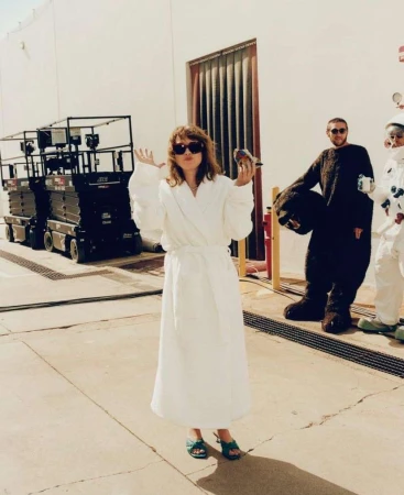Woman in white robe poses with a bird, two people behind her in an industrial setting with aerial lifts and a closed door.