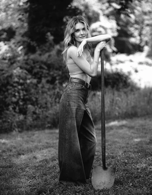 Black and white shot of a woman with long hair posing outdoors with a shovel, wearing a tank top and wide-leg jeans.