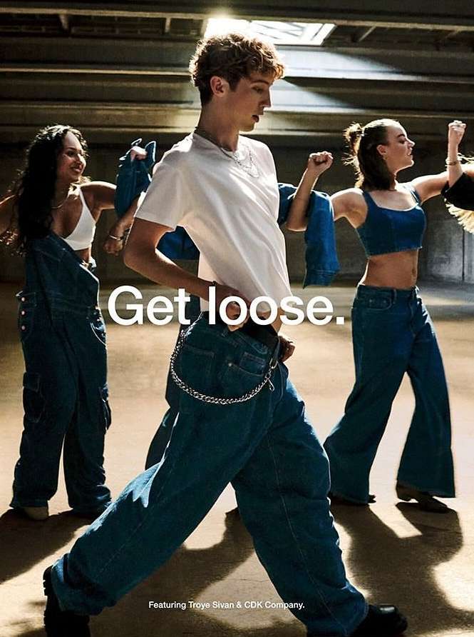 Three people wearing oversized denim clothing pose indoors, with the central figure in a white t-shirt and jeans with a chain.