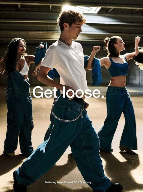 Three people wearing oversized denim clothing pose indoors, with the central figure in a white t-shirt and jeans with a chain.