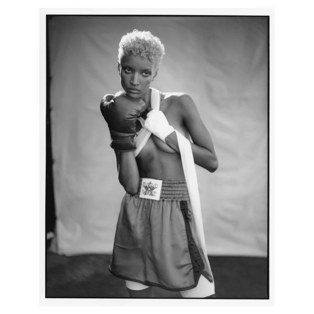 Black and white studio shot of a confident female boxer with boxing gloves, wrapped hands, and shorts against a simple backdrop.