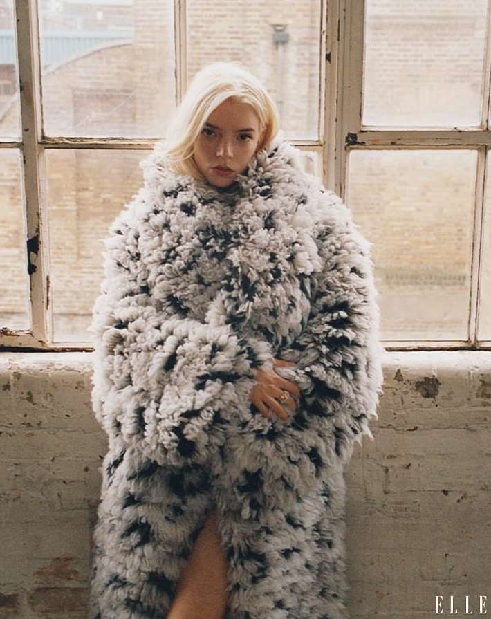 Fashion portrait of Anya Taylor-Joy wearing a long, textured coat next to a window, showcasing a blend of textures and neutral tones.