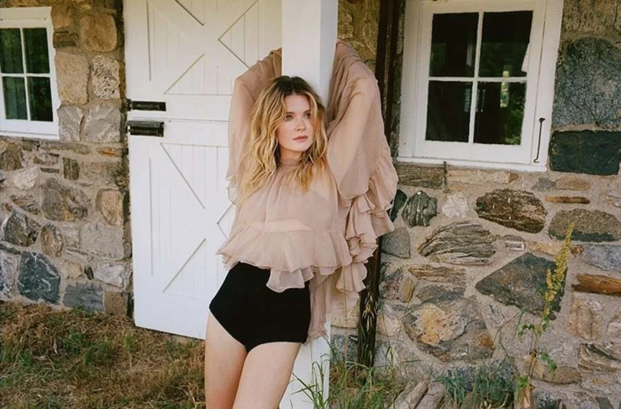 Woman with wavy blonde hair poses in a ruffled top and shorts near a stone building with white doors and windows outdoors.