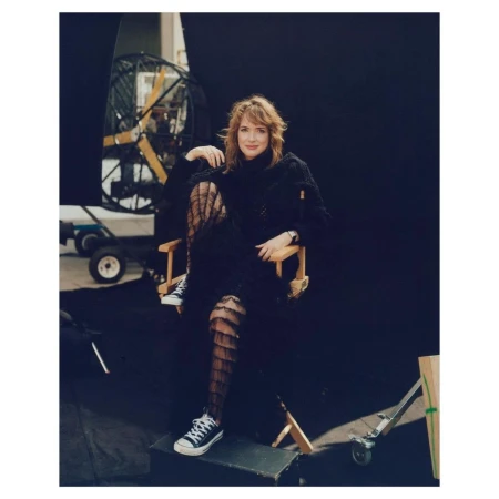A woman sits in a director's chair on a film set wearing a black outfit and sneakers, with film equipment visible in the background.