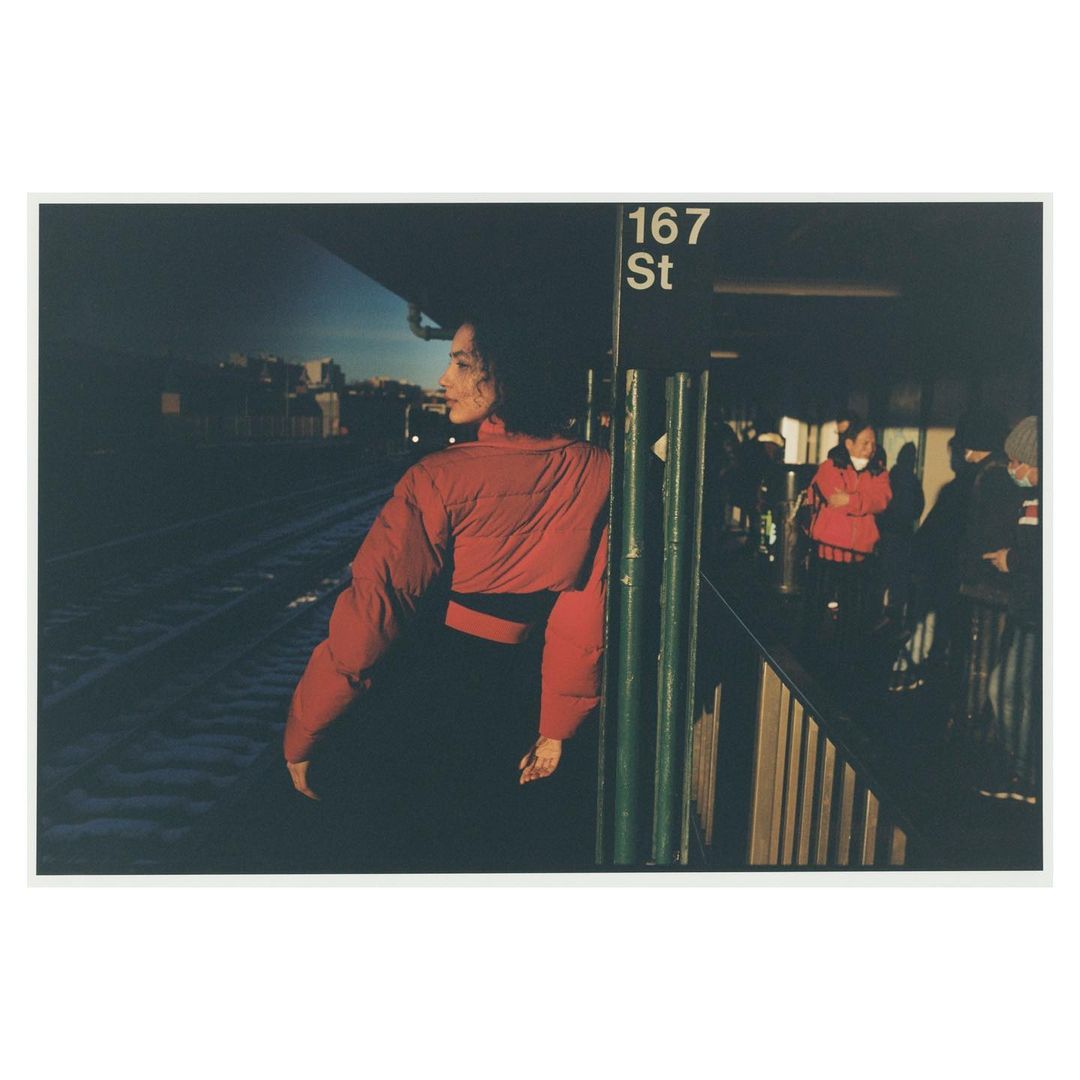 Woman in red jacket waits at outdoor subway station in urban environment with several other people in the background.