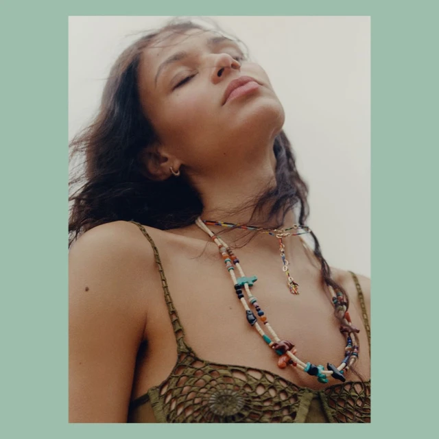 Young woman with eyes closed wearing colorful beaded necklaces in an outdoor setting, looking upward in a portrait style.