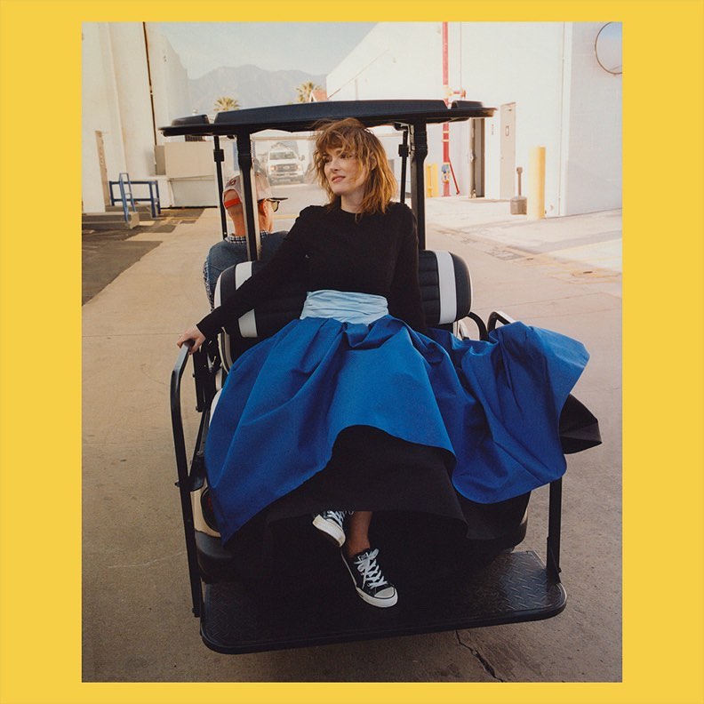 A woman in a blue gown sitting in a golf cart with an adult male in the background, captured on a sunny day in an urban setting.
