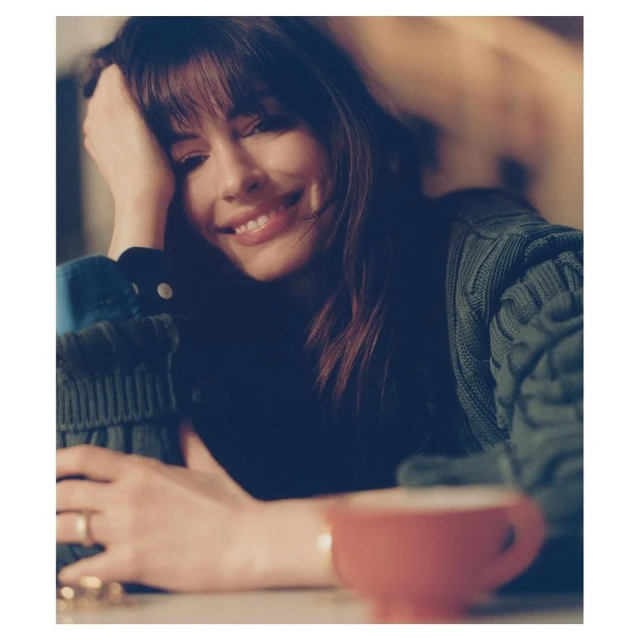 A woman with bangs is smiling while leaning on her hand. She is wearing a green cable-knit sweater and has a cup in front of her.