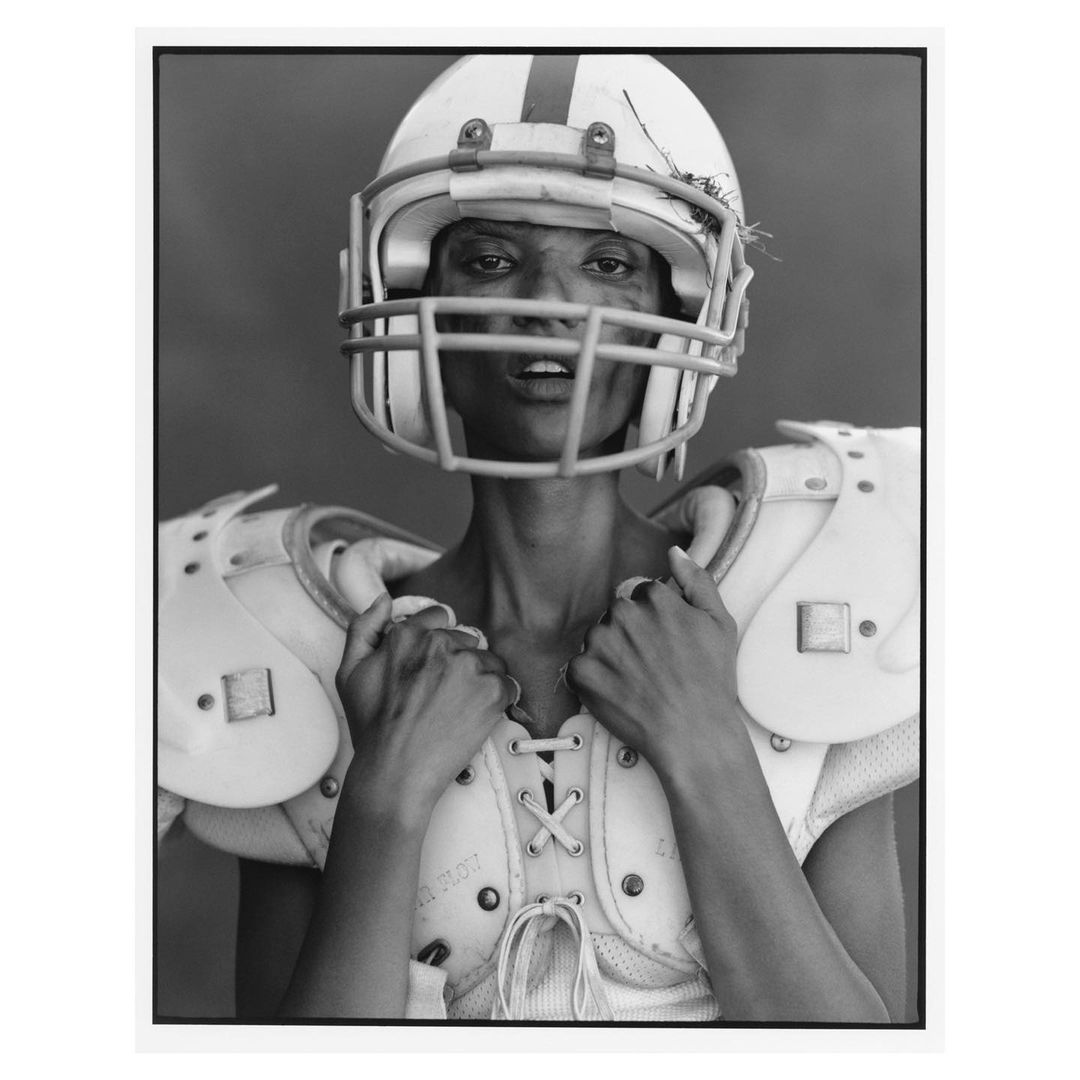 Black and white studio portrait of a woman in football gear including shoulder pads and a helmet, standing against a grey background.
