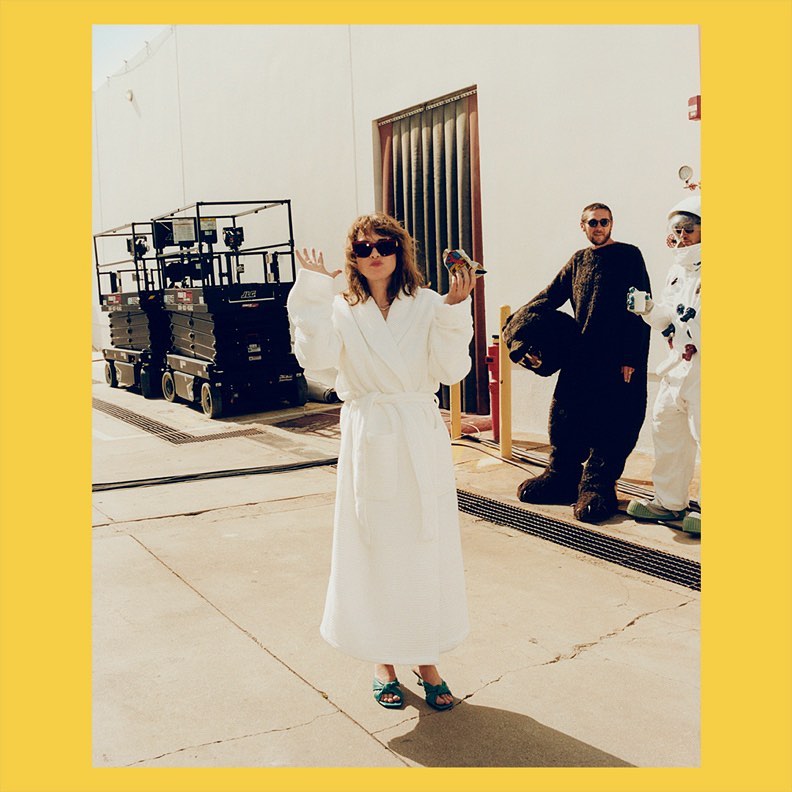 Woman in a white robe stands in front of industrial equipment with two other people, one in a bear suit and another in a white suit.