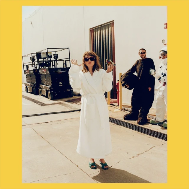 Woman in a white robe stands in front of industrial equipment with two other people, one in a bear suit and another in a white suit.