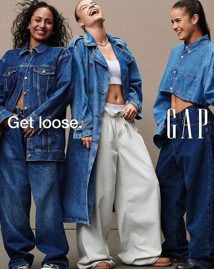 Three smiling women pose in denim jackets and blue and white wide-leg pants for a GAP fashion advertisement against a neutral backdrop.