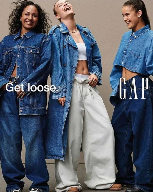Three smiling women pose in denim jackets and blue and white wide-leg pants for a GAP fashion advertisement against a neutral backdrop.