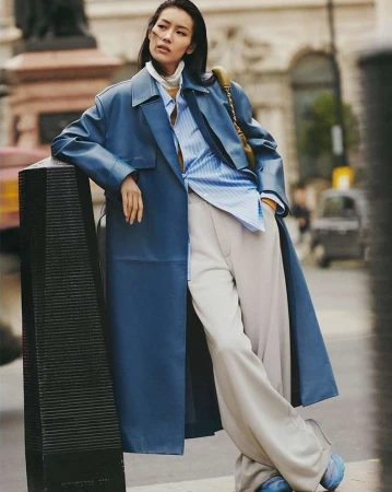 Model in blue leather coat and pinstripe shirt leaning against a bollard on a London street