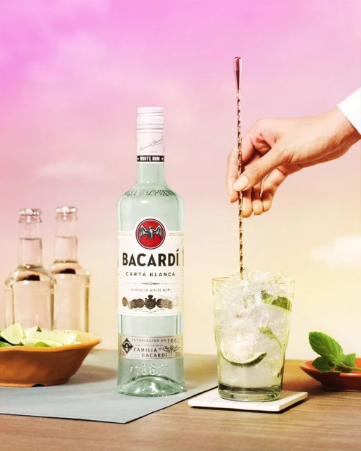 Bacardi Carta Blanca white rum is stirred into a cocktail with limes and mint, set against a soft pink and orange gradient background.