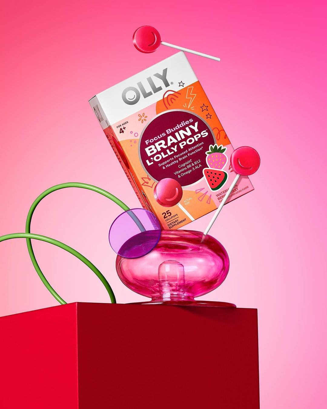 Box of Olly Brainy L'Olly Pops dietary supplement with lollipops, transparent pedestal, and red cube against a pink background.
