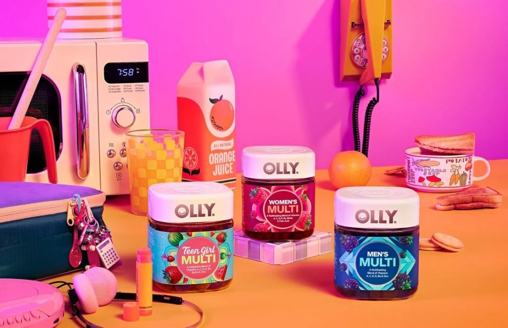 Eye-level shot of three Olly vitamin containers, an orange juice box, a microwave, and a retro yellow phone on an orange surface.