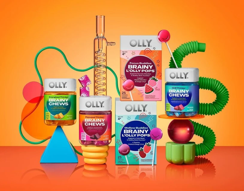 Still life of Olly Brainy Chews and L'Olly Pops vitamin supplements in colorful packaging, arranged with geometric shapes on orange backdrop.