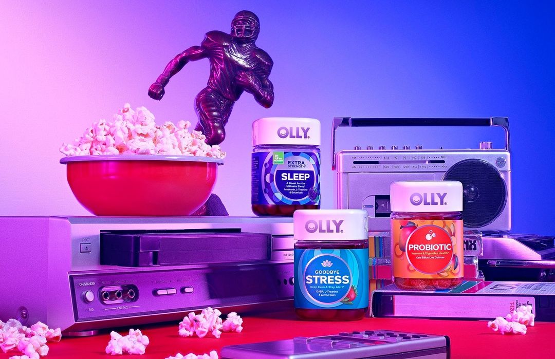 Olly sleep and probiotic supplement bottles sit among retro electronics, popcorn, and a football trophy under vibrant neon lighting.