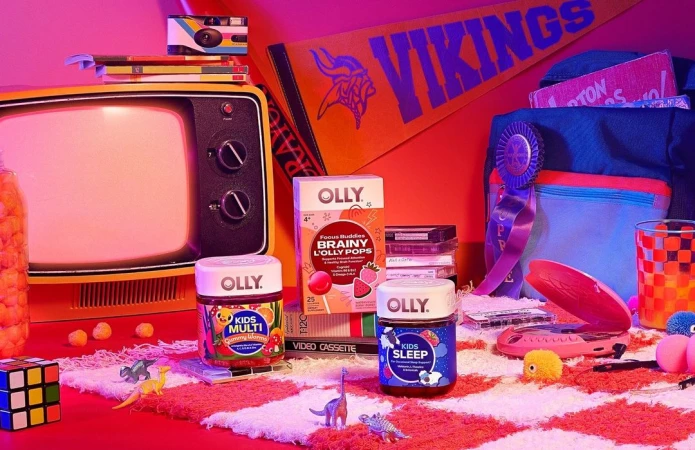 Nostalgic 80s-themed product shot of Olly kids vitamins with a vintage TV, VHS tapes, pennant, and Rubik's cube against pink backdrop.