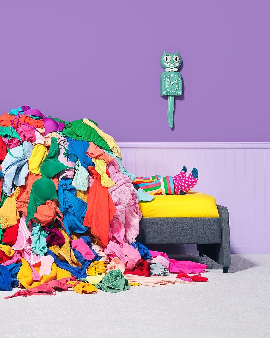 Person lies on a bed half-buried under a mountain of brightly colored clothes with a quirky cat clock on a purple wall.