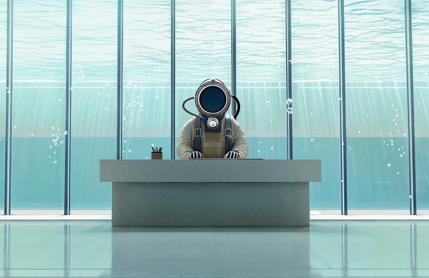 Illustration of a person in a diving suit sitting at a desk with office supplies. The setting is an underwater office with sunlight.