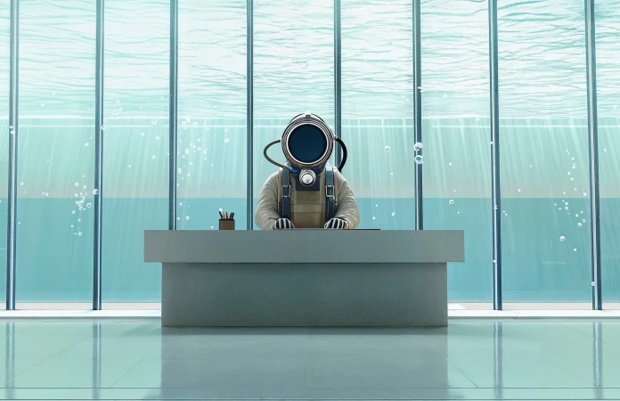 Illustration of a person in a diving suit sitting at a desk with office supplies. The setting is an underwater office with sunlight.