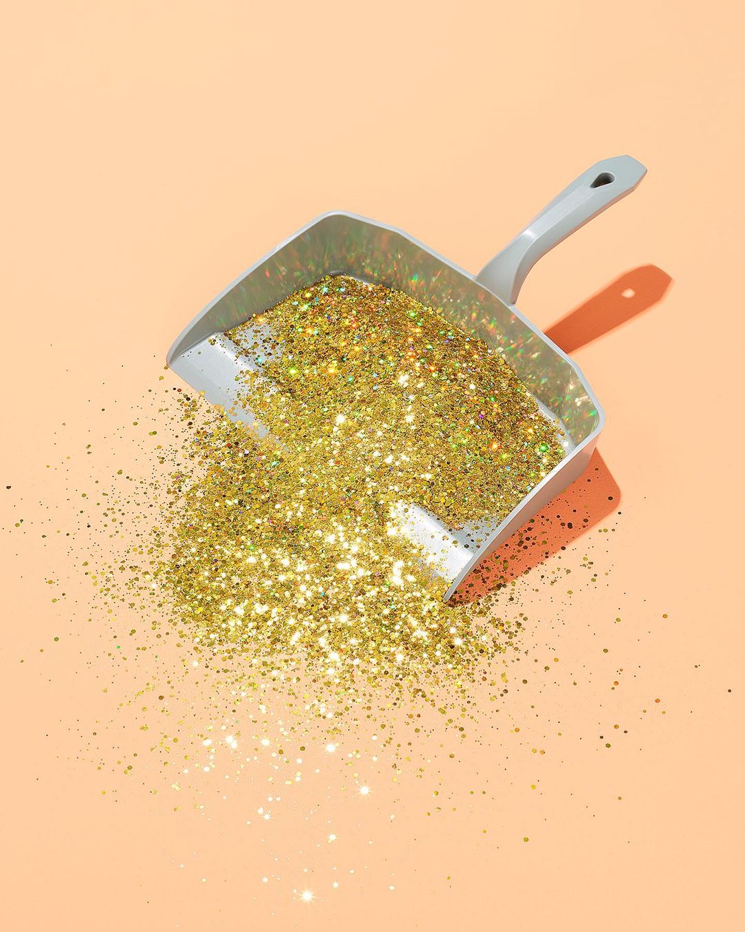 Dustpan full of gold glitter on a peach-colored background with direct lighting creating a defined shadow from the object.