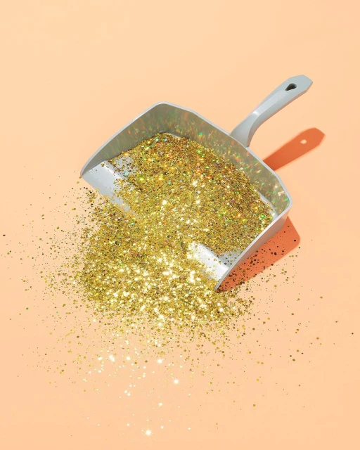 Dustpan full of gold glitter on a peach-colored background with direct lighting creating a defined shadow from the object.