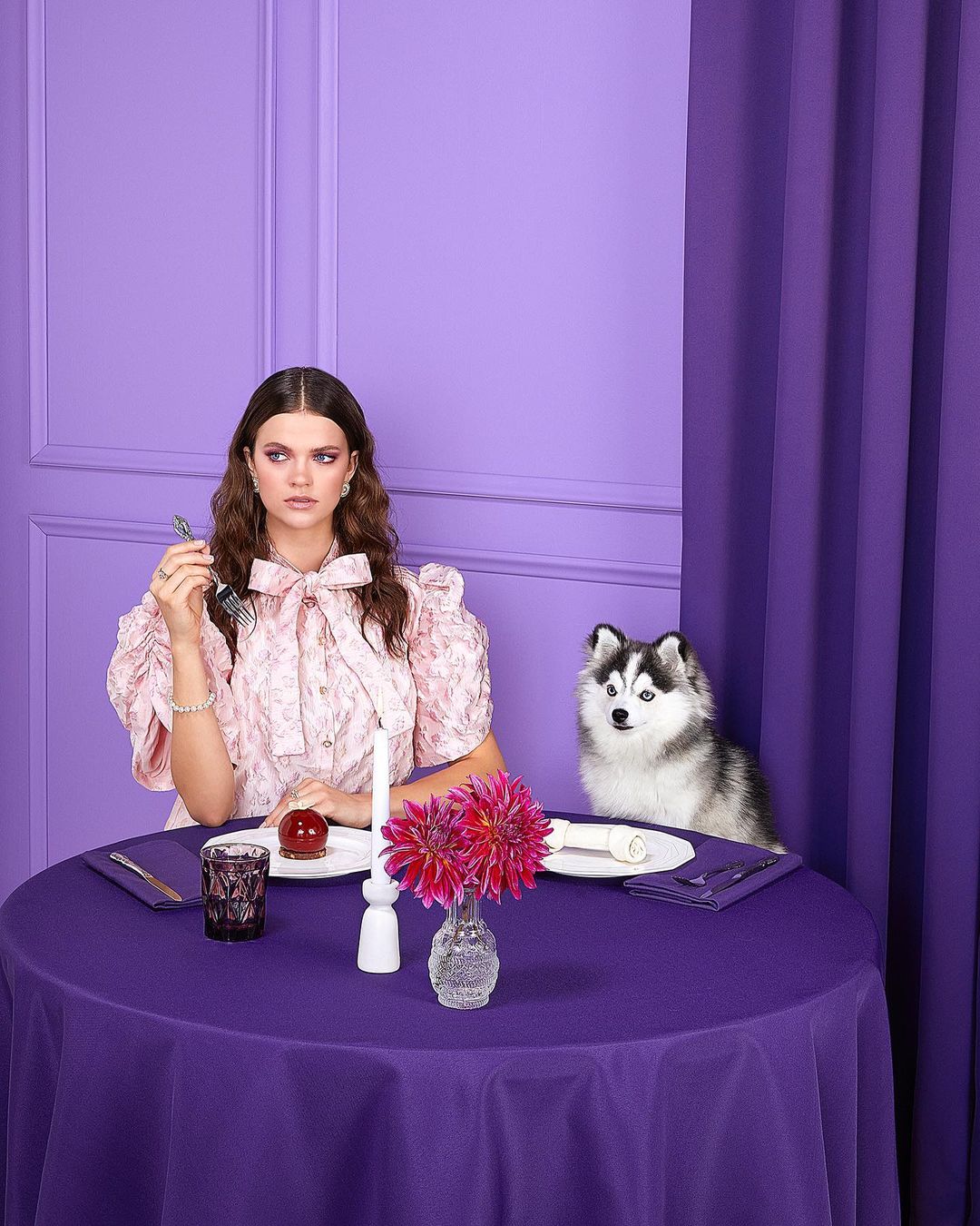 Elegant woman with a husky at a table set with a purple tablecloth, candle, and a vase of pink flowers against a lilac backdrop.