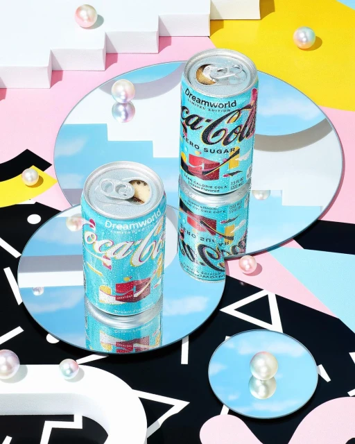 Two cans of Coca-Cola Dreamworld Zero Sugar on reflective surfaces, surrounded by white geometric shapes and pearl beads.
