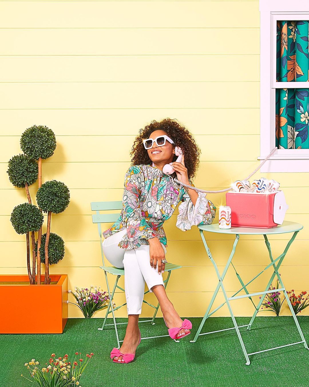 A woman with curly hair and white sunglasses talks on a pink retro telephone while sitting on a light green chair at a table.