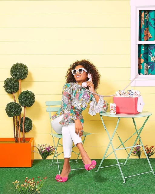 A woman with curly hair and white sunglasses talks on a pink retro telephone while sitting on a light green chair at a table.