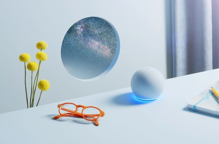 Still life of a white spherical smart speaker with blue light, orange glasses, yellow flowers, and a notebook on a blue table indoors.