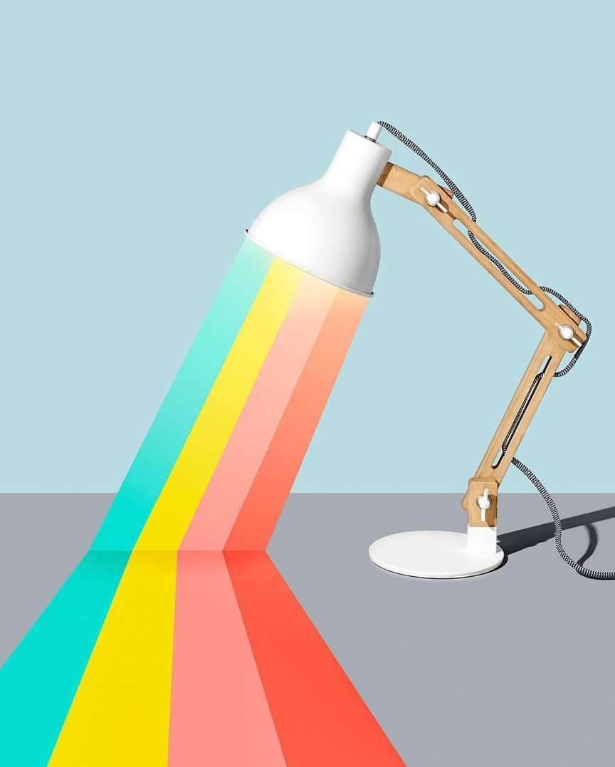 Studio shot of a desk lamp with wooden arm emitting rainbow-colored light on a gray surface against a light blue background.
