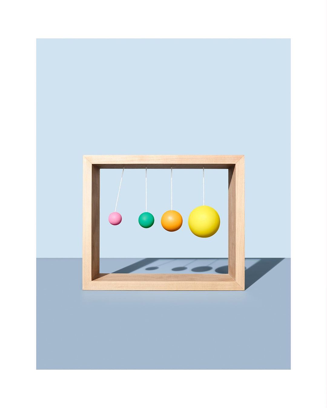 Kinetic art piece featuring colorful spheres suspended in a wooden frame, casting shadows against a light blue and gray background.