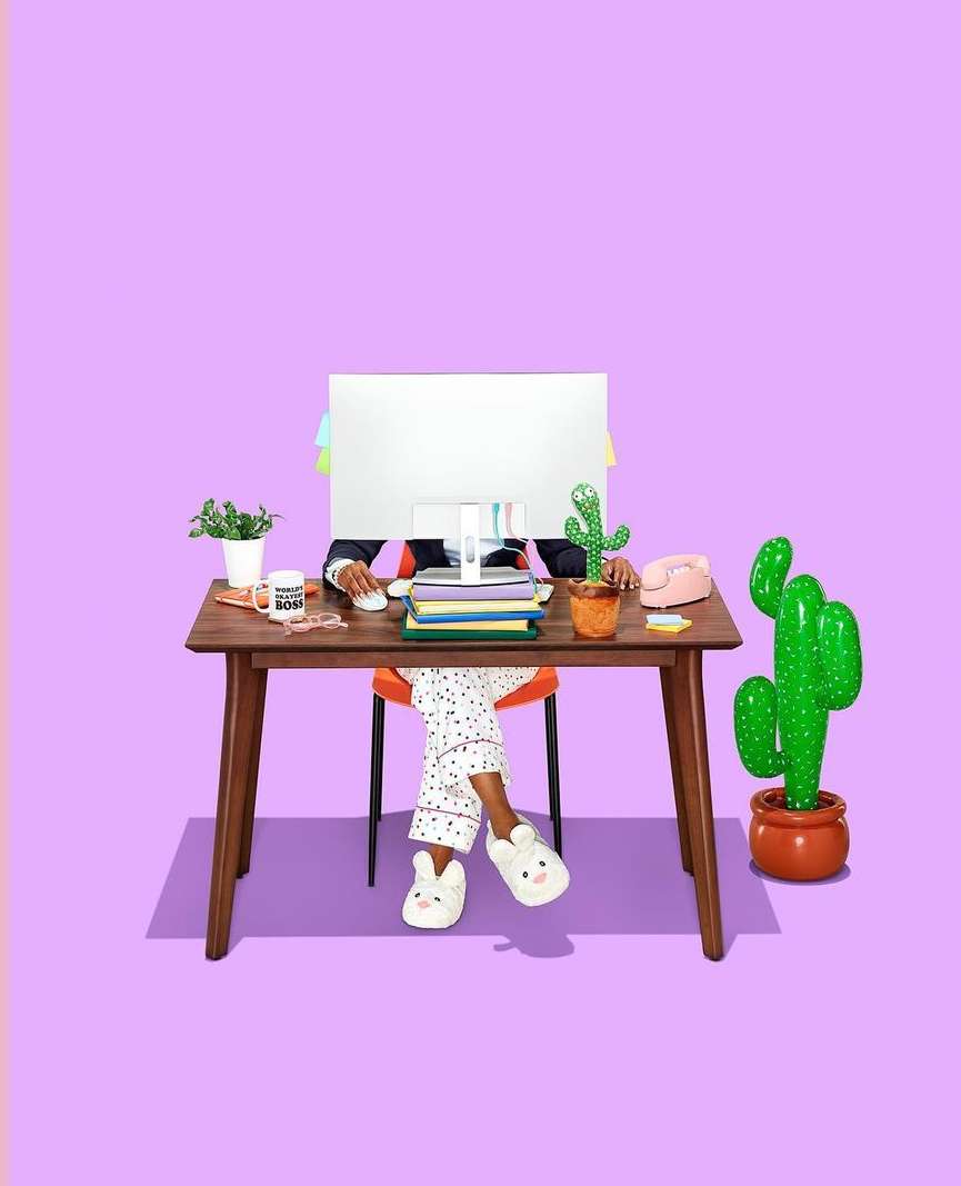 Person wearing pajama pants and slippers at a desk working on a computer in a studio with a lilac background and cactus.