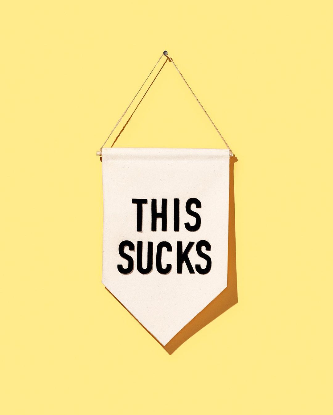 A white pennant with the text 'THIS SUCKS' in black letters hangs against a light yellow wall, casting a soft shadow.