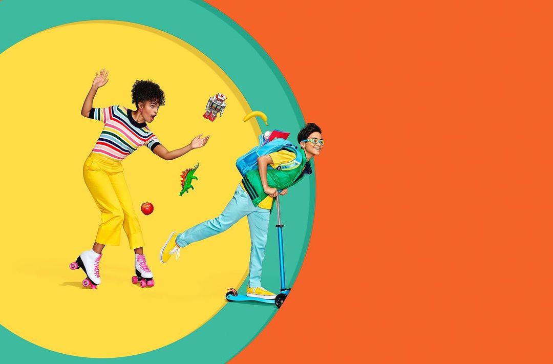 Playful studio shot featuring a young woman on roller skates with a rainbow shirt and a child riding a scooter on a yellow and orange background.