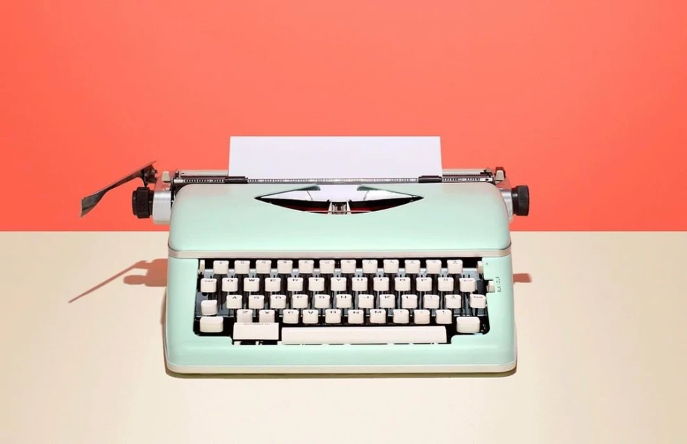 Close-up of a mint-green vintage typewriter on a tan surface against a salmon-colored background, showcasing its retro design and details.