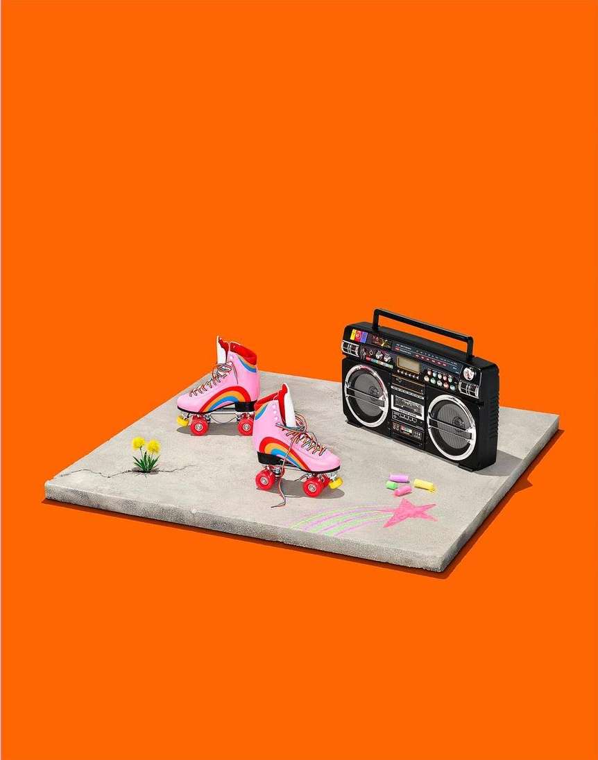 A pair of pink roller skates with rainbows next to a black boombox on a grey platform against a solid orange backdrop in a studio setting.