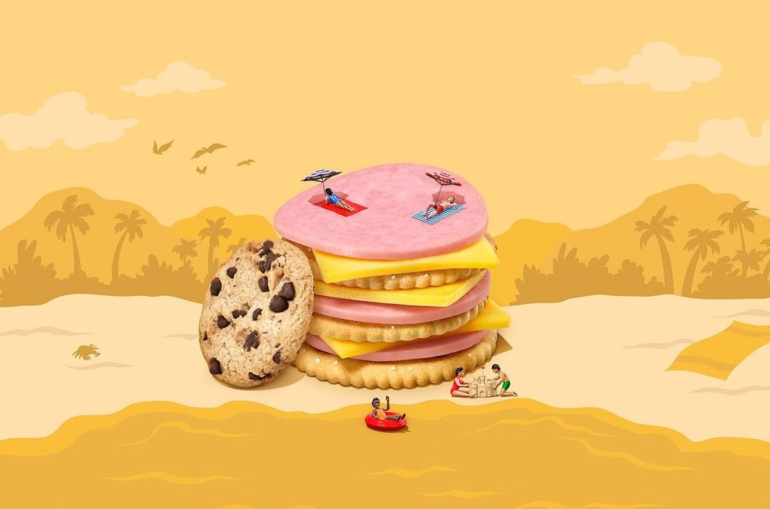 A whimsical food scene with a giant sandwich-cookie stack and miniature people relaxing on the layers against a soft yellow beach backdrop.