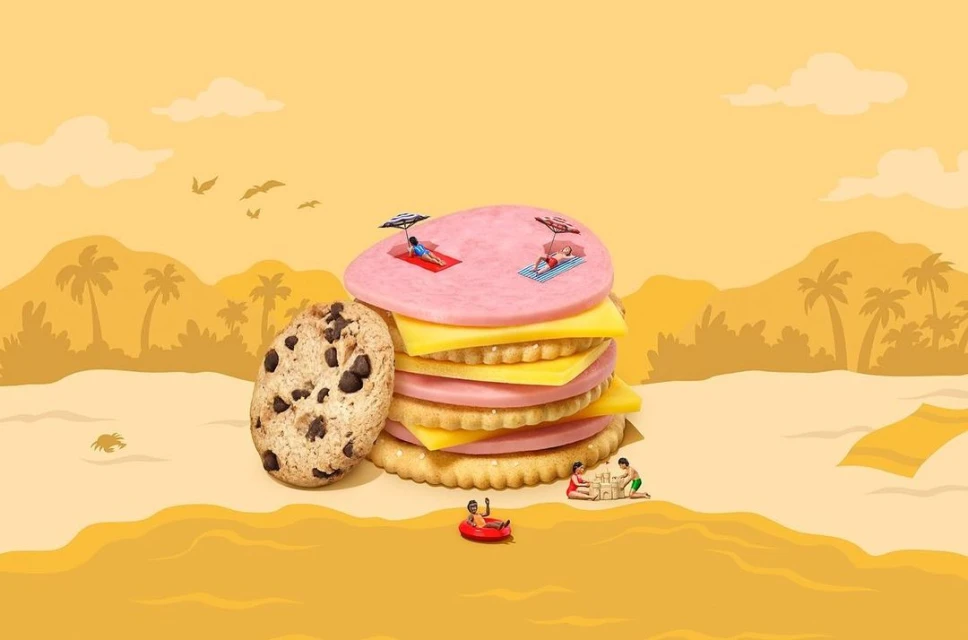 A whimsical food scene with a giant sandwich-cookie stack and miniature people relaxing on the layers against a soft yellow beach backdrop.