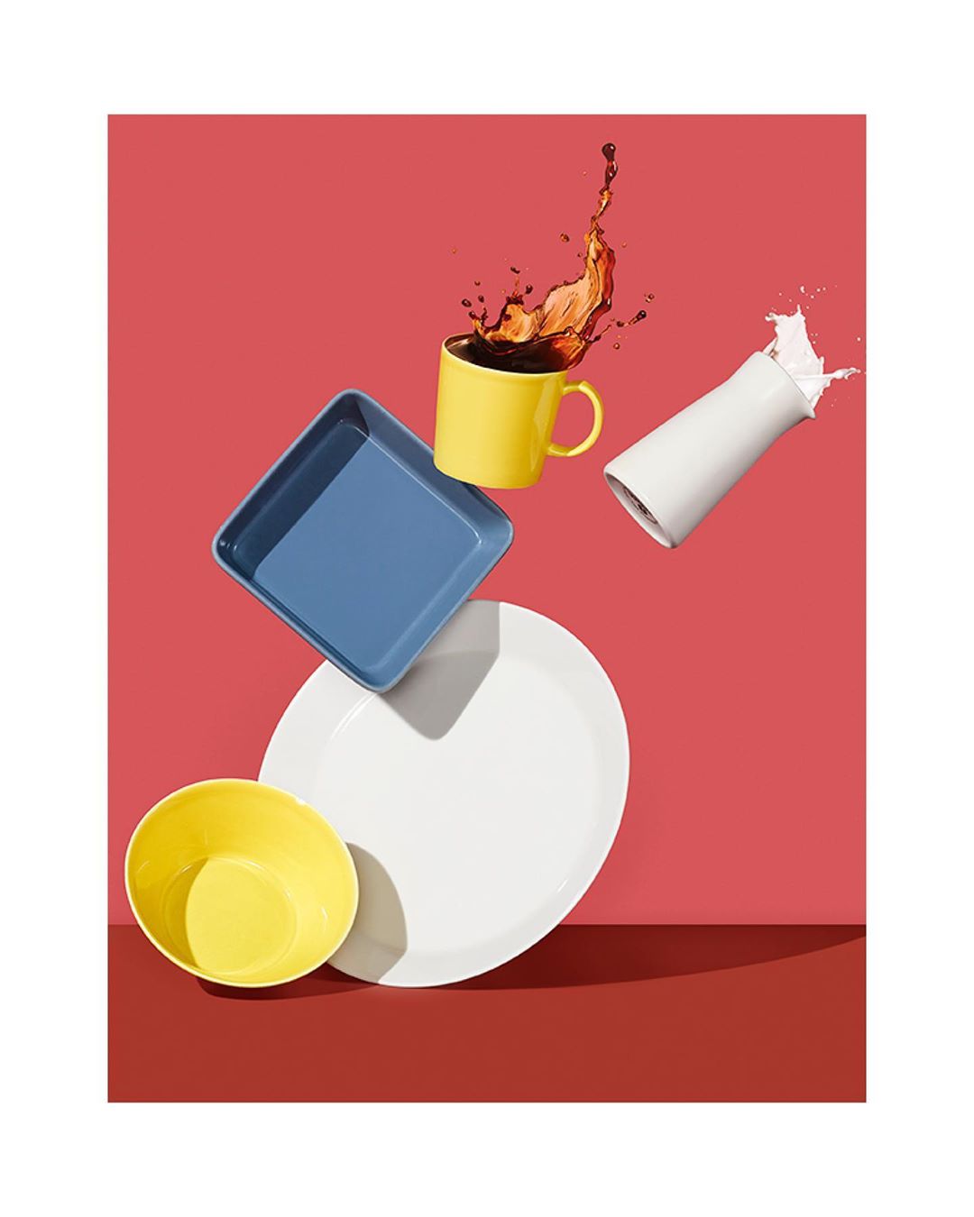 Elevated view of a white plate, blue square baking dish, yellow bowl, and mug with coffee and milk splashing on a coral surface.