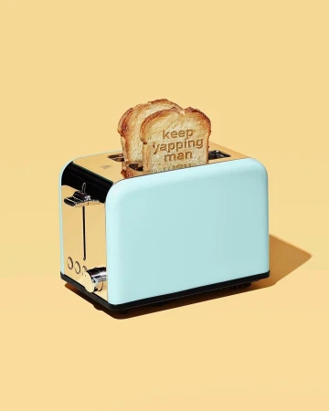 A light-blue and gold toaster features two slices of toast with stamped letters spelling out 'keep yapping man' on a yellow background.