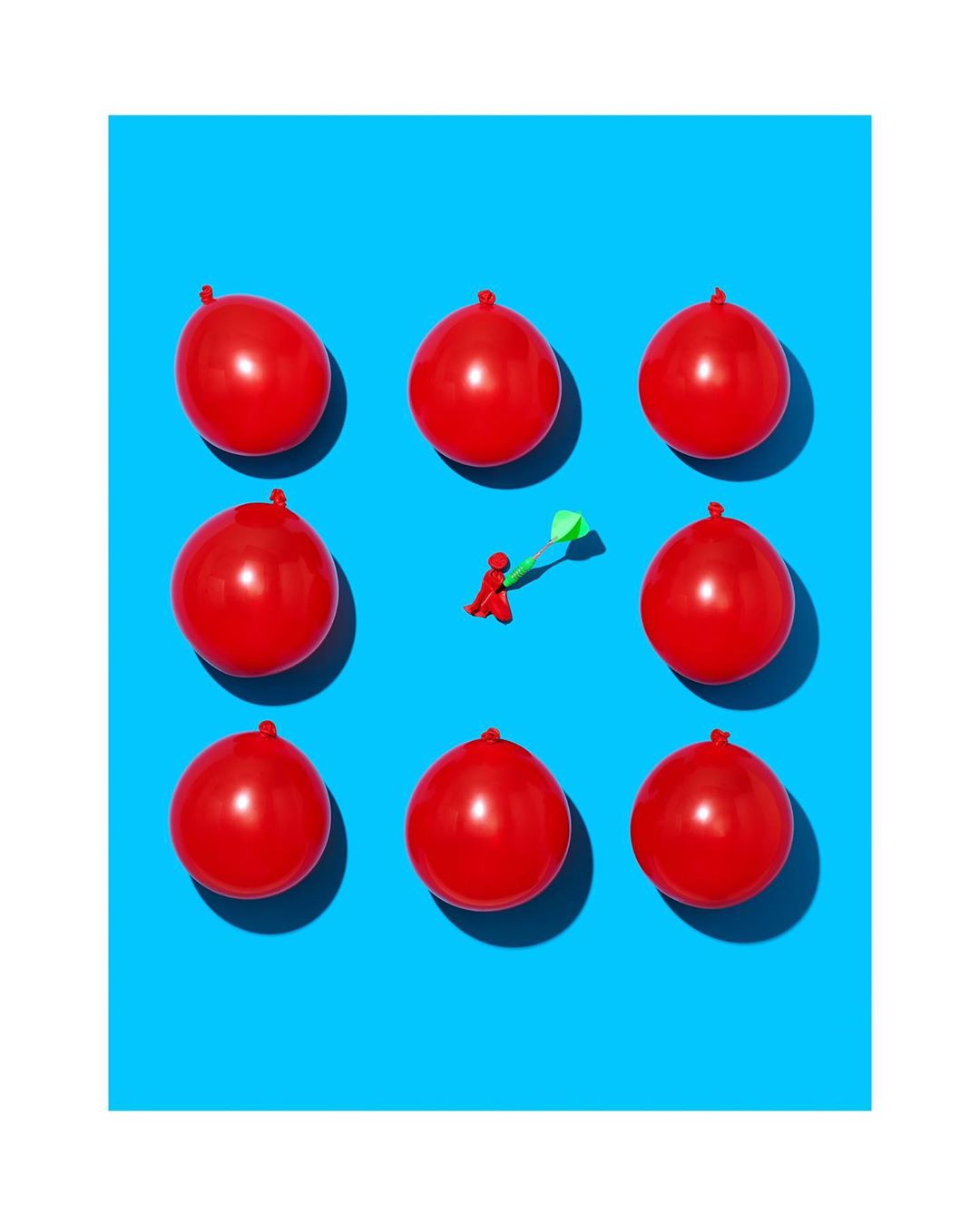 Conceptual image featuring red balloons arranged in a grid with a red figure throwing a dart on a blue background, studio shot.