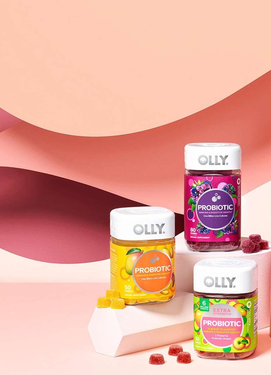 Three bottles of Olly Probiotic Gummies with peach, blueberry, and mango flavors displayed on a pink geometric surface.